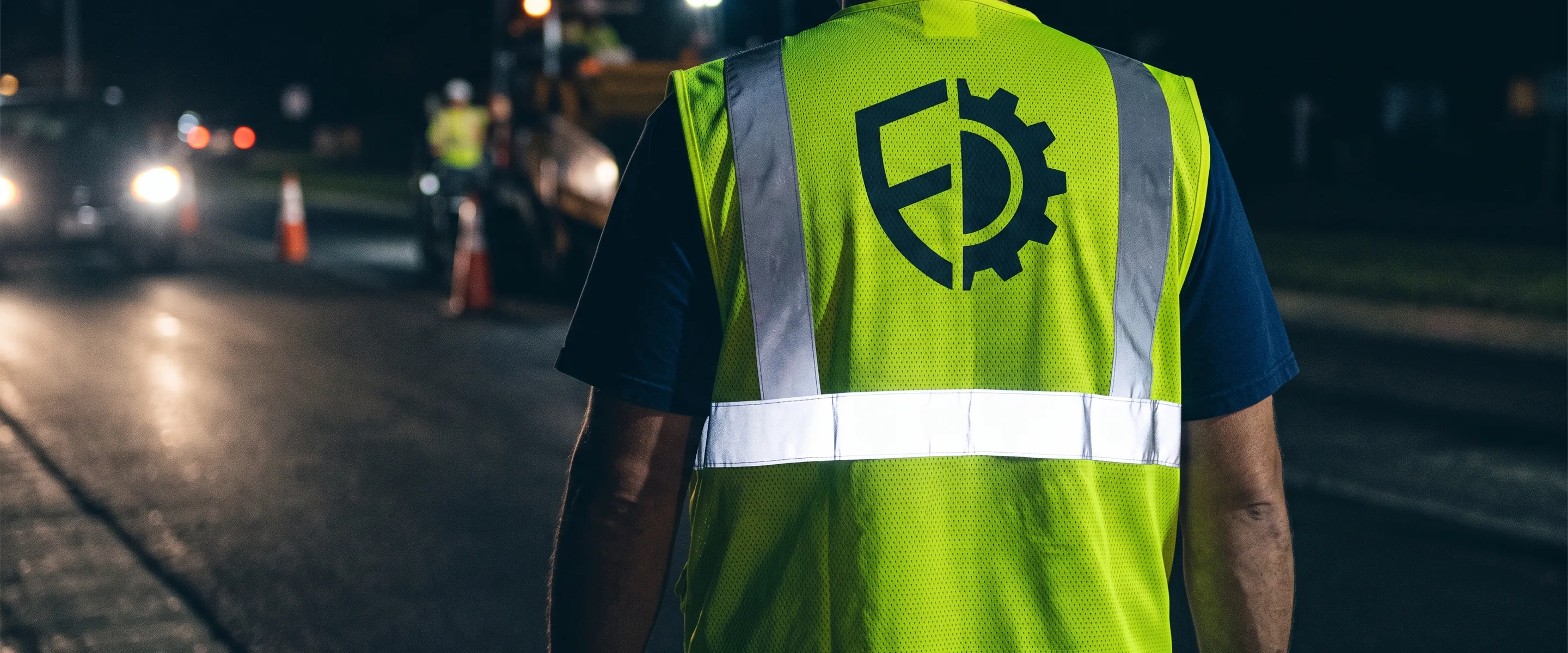 Back of a safety vest with a large logo above the horizontal reflective tape, lit by vehicle headlights at night