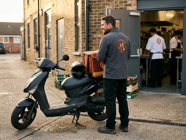 Delivery rider in a softshell jacket with a logo on the back beside a moped